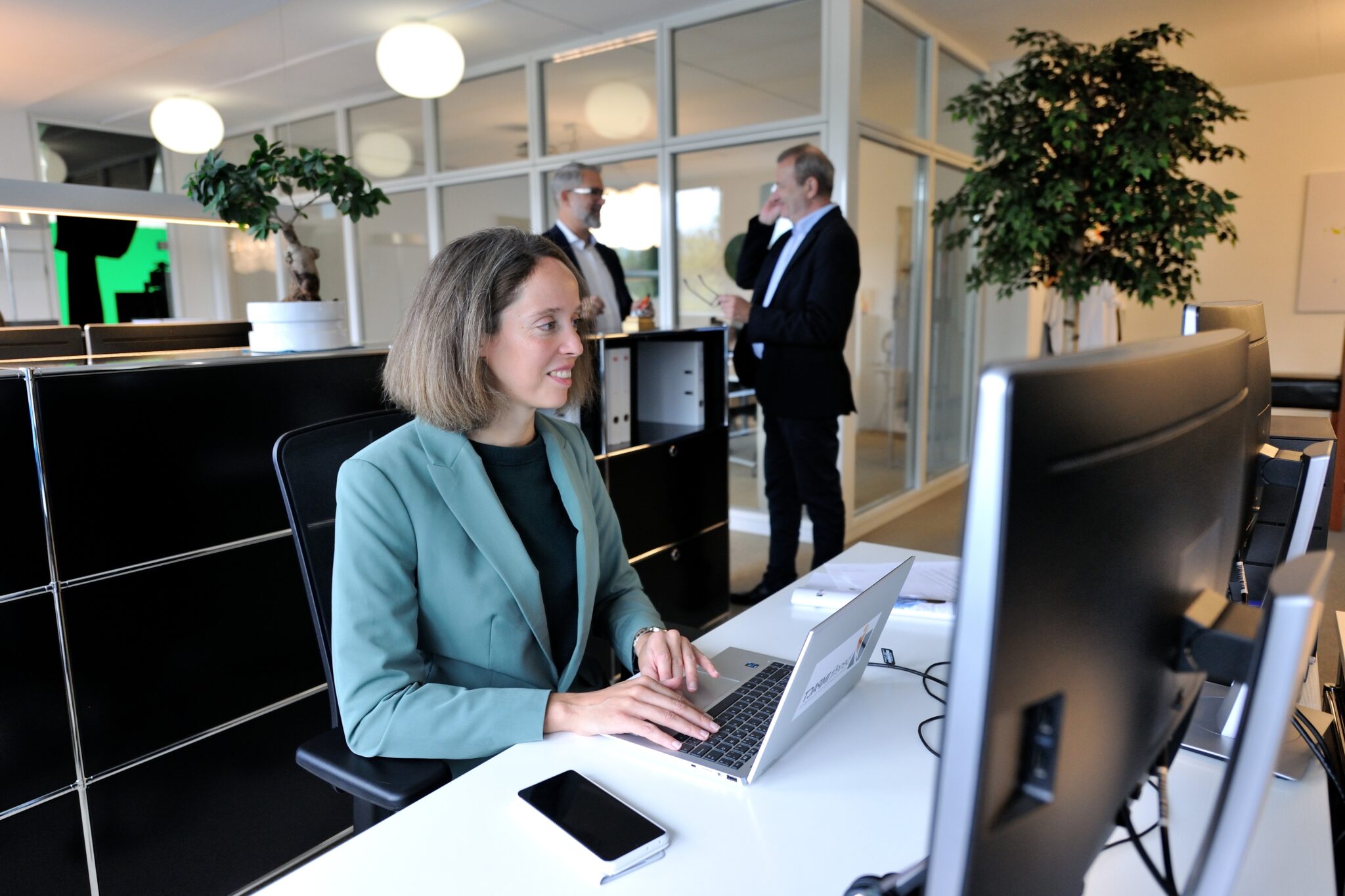 benefitIMPACT Team im Arbeitsalltag - Projekt- & Officemanagerin Luce Vormoor am Laptop, im Hintergrund Inhaber Pascal Yves SChwartz im Gespräch mit Freelancer & Schauspieler Jean-Christophe Nigon